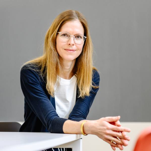 Dr. Isabell Osann, Coach im Coaching Kontext, An einem Tisch sitzend, ruhig blickend, mit einer Tafel im Hintergrund.